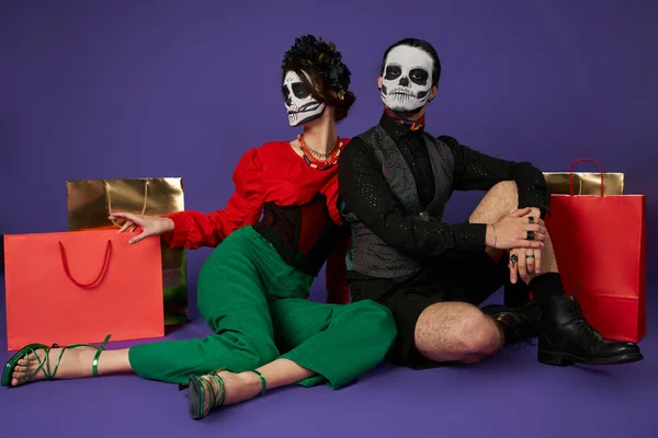 Stylish couple in dia de los muertos skull makeup sitting near shopping bags on blue, full length — Stock Photo