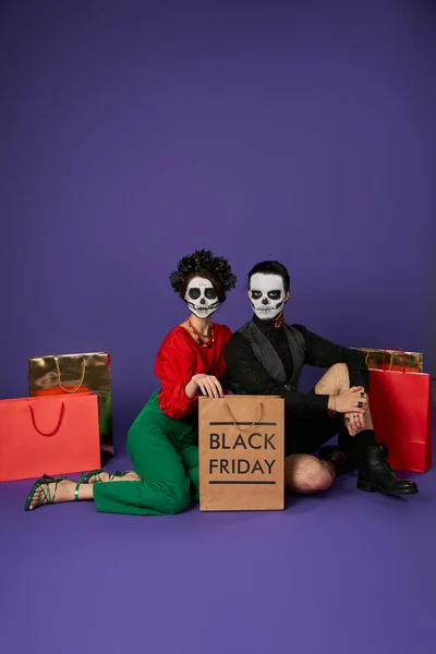 Casal na moda na maquiagem crânio dia de los muertos sentado perto de sacos de compras na sexta-feira azul, preto — Fotografia de Stock