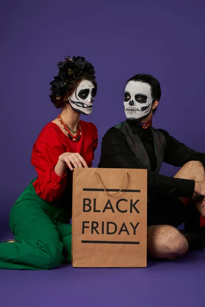 Giovane coppia in tradizionale dia de los muertos trucco seduto vicino nero venerdì shopping bag su blu — Foto stock