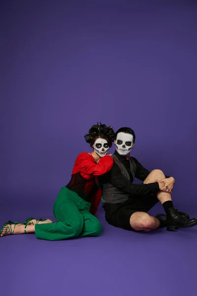 Couple dans le traditionnel dia de los muertos maquillage et tenue de fête assis sur bleu, pleine longueur — Photo de stock