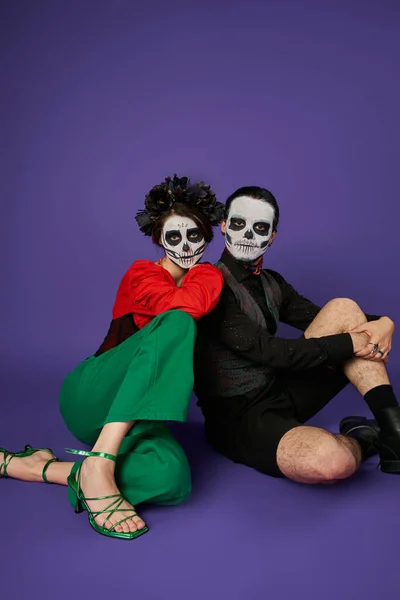 Dia de los muertos couple, woman in skull makeup leaning on spooky man while sitting on blue — Stock Photo