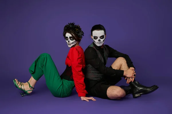 Paar in Zuckerschädel-Make-up und trendigem Outfit Rücken an Rücken auf blau sitzend, dia de los muertos fest — Stockfoto