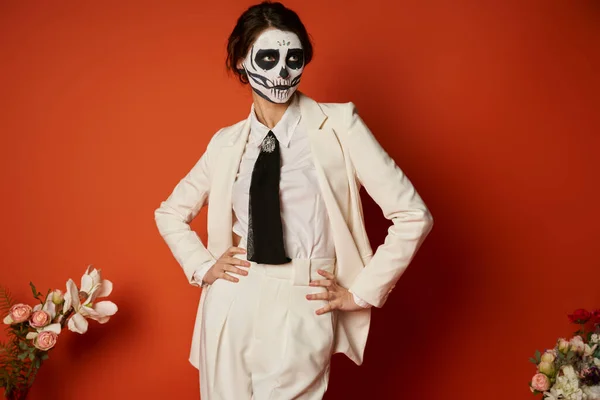 Woman in skeleton makeup and white suit posing with hands on hips near flowers on red, Day of Dead — Stock Photo