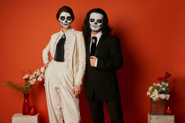 Elegant couple in skull makeup and suits near altar with flowers on red, near dia de los muertos — Stock Photo