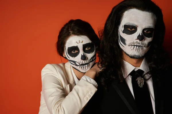 Elegante pareja de día de los muertos en maquillaje de cráneo, mujer apoyada en el hombro del aterrador hombre sobre rojo - foto de stock