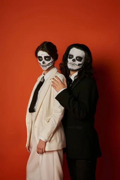 Elegant couple in dia de los muertos makeup and elegant festive suits looking at camera on red — Stock Photo