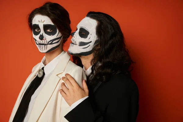 Stylish couple in dia de los muertos makeup, spooky man hugging woman looking at camera on red — Stock Photo
