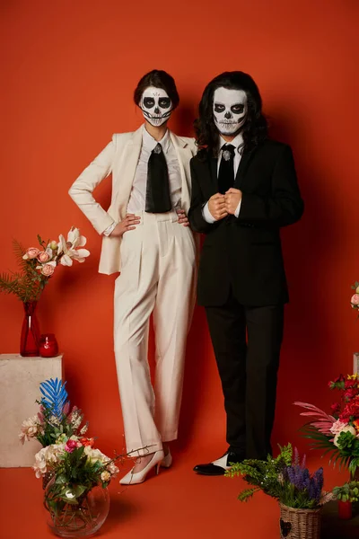 Couple in scary skull makeup and suits near festive dia de los muertos ofrenda with flowers on red — Stock Photo