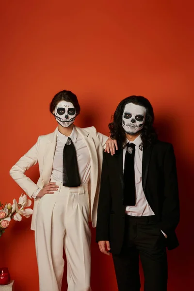 Stylish couple in skull makeup and elegant suits posing near flowers and looking at camera on red — Stock Photo