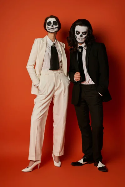 Full length of stylish couple in skull makeup and elegant suits posing with hands in pockets on red — Stock Photo