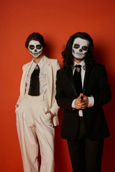 Modisches Paar im Totenkopf-Make-up, Frau mit Händen in den Taschen, die in die Kamera schaut, in der Nähe eines beängstigenden Mannes auf Rot — Stockfoto