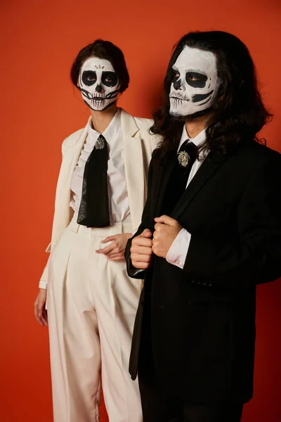 Woman in catrina makeup and white suit looking at camera near scary man on red, dia de lost muertos — Stock Photo