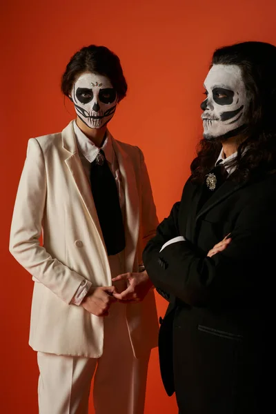 Paar in dia de los muertos Make-up, Frau im weißen Anzug in der Nähe beängstigender Mann mit gefalteten Armen auf rot — Stockfoto