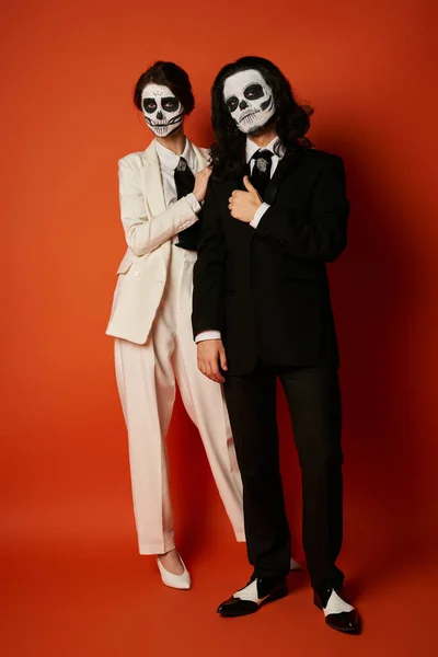 Full length of couple in dia de los muertos skull makeup and festive suits looking at camera on red — Stock Photo