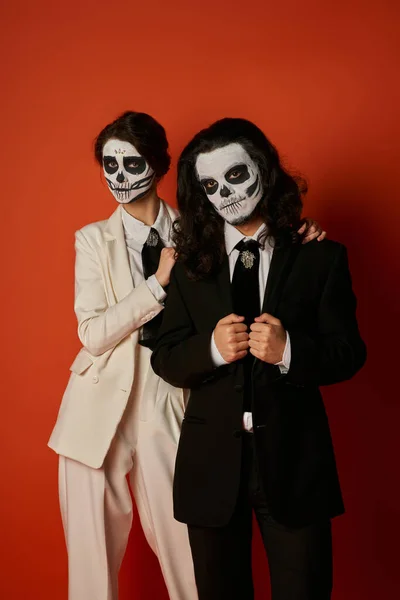 Elegante pareja en día de los muertos azúcar cráneo maquillaje y trajes mirando a la cámara en el fondo rojo - foto de stock