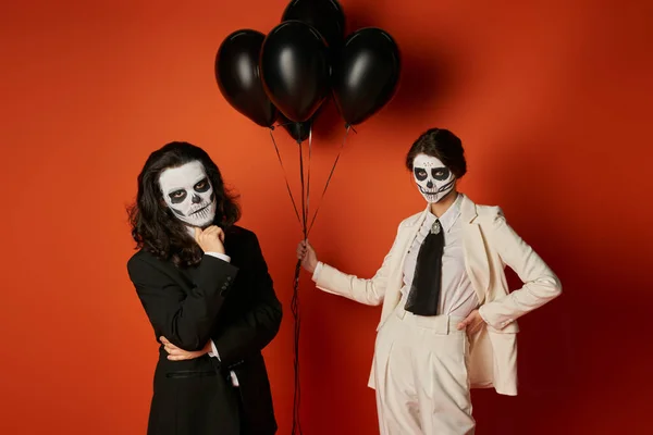 Mulher em maquiagem crânio e terno branco com balões pretos perto de homem assustador no vermelho, dia de los muertos — Fotografia de Stock