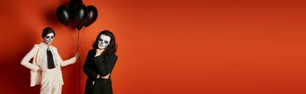 Femme dans le maquillage du crâne de sucre et costume blanc avec des ballons noirs près homme effrayant sur rouge, bannière — Photo de stock