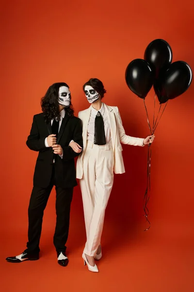 Пара в котячому макіяжі і костюмах дивиться один на одного біля чорних кульок на червоному, День мертвих — стокове фото