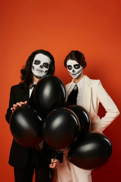 Fiesta de día de los muertos, pareja espeluznante en maquillaje de cráneo mirando a la cámara cerca de globos negros sobre rojo - foto de stock