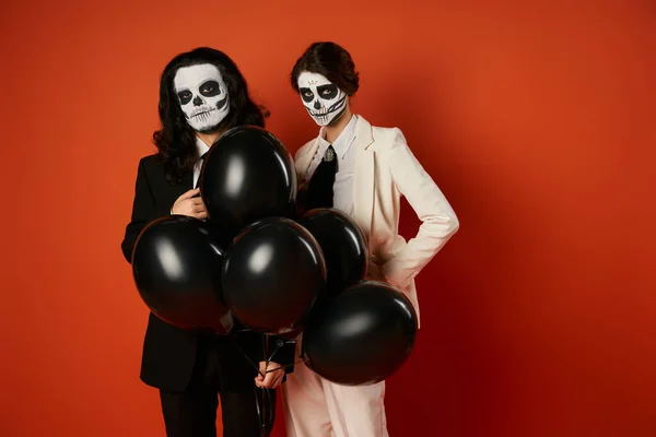 Eerie couple in catrina makeup and suits posing with black balloons on red, dia de los muertos party — Stock Photo