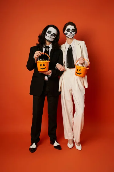 Toute la longueur du couple élégant dans le maquillage catrina tenant des seaux de bonbons sur le rouge, Jour de la fête des morts — Photo de stock