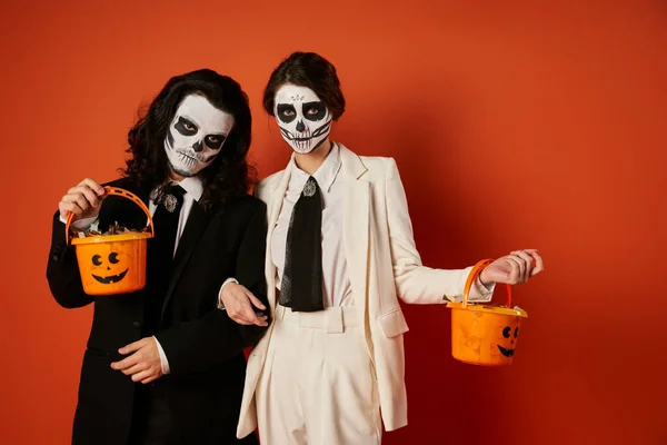 Elegante casal no dia de los muertos maquiagem segurando baldes de doces e olhando para a câmera no vermelho — Fotografia de Stock