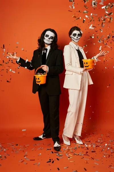 Pareja en maquillaje cráneo lanzando confeti brillante de baldes de caramelo en rojo, fiesta de día de los muertos - foto de stock