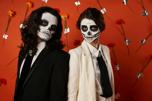 Trendy couple in dia de los muertos skull makeup looking at camera on red backdrop with flowers — Stock Photo