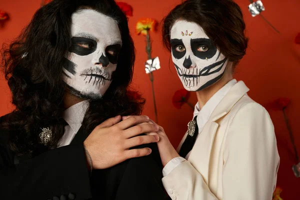 Woman in dia de los muertos skull makeup touching shoulder of man on red backdrop with carnations — Stock Photo