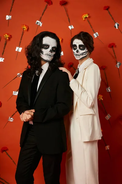 Woman in dia de los muertos catrina makeup touching shoulder of man on red backdrop with carnations — Stock Photo