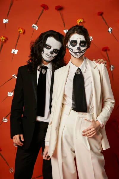 Couple in dia de los muertos skull makeup and suits looking at camera on red backdrop with flowers — Stock Photo