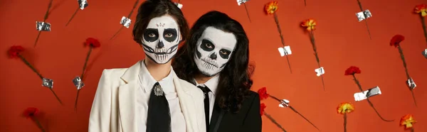 Couple in sugar skull makeup and suits looking at camera on red backdrop with carnations, banner — Stock Photo