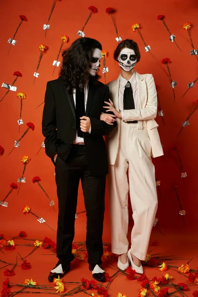 Longitud completa de la pareja en el maquillaje catrina y traje festivo posando sobre fondo rojo con claveles - foto de stock