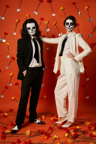 Full length of stylish couple in skull makeup and suits standing on red backdrop with carnations — Stock Photo