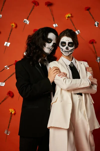 Dia de los muertos couple, woman with folded arms near spooky man on red backdrop with carnations — Stock Photo
