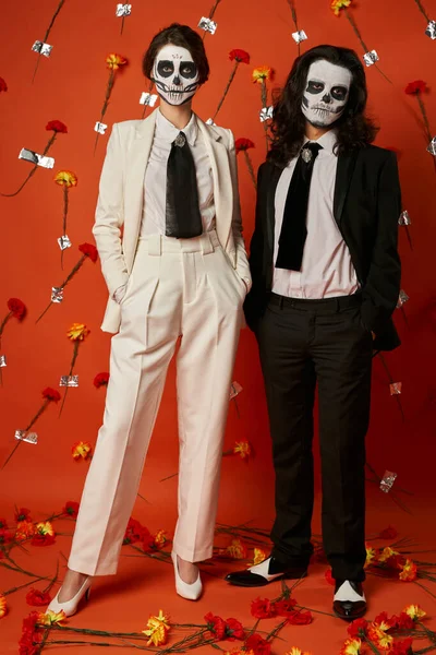 Couple dans le maquillage du crâne de sucre et tenue tendance posant avec les mains dans des poches de fond floral rouge — Photo de stock