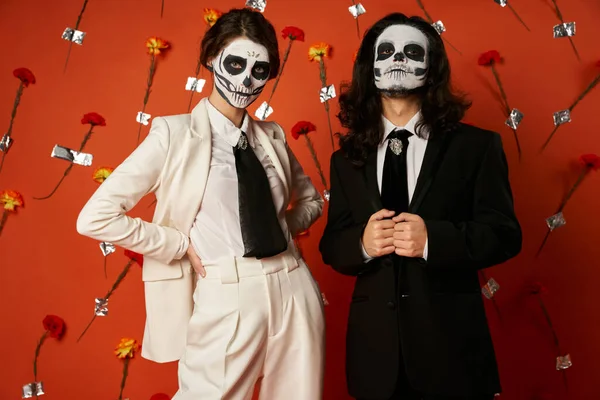Trendy couple in sugar skull makeup and suits looking at camera on red backdrop with carnations — Stock Photo
