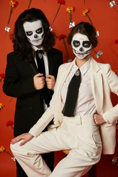 Elegantes Paar in Catrina-Make-up und Anzügen posiert auf rotem Hintergrund mit Nelken, Day of Dead — Stockfoto
