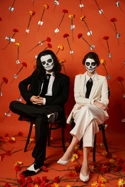 Пара в костюмах для макияжа черепа сидит на стульях в красной студии с цветочным декором, dia de los muertos — стоковое фото