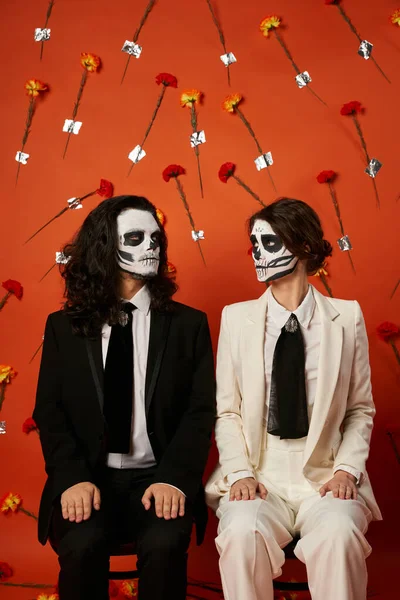 Espeluznante pareja de día de los muertos sentados en sillas y mirándose el uno al otro sobre fondo floral rojo - foto de stock