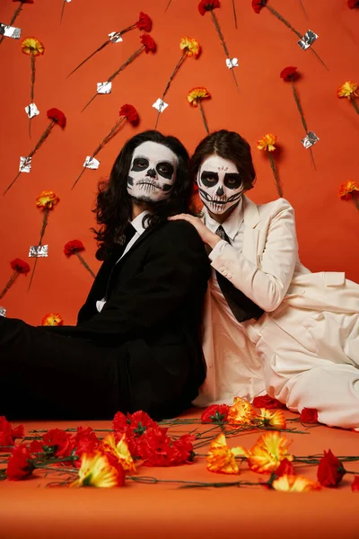 Elegantes Paar in Zuckerschädel-Make-up und Anzügen sitzt auf dem Boden in rotem Studio mit Nelkenblüten — Stockfoto