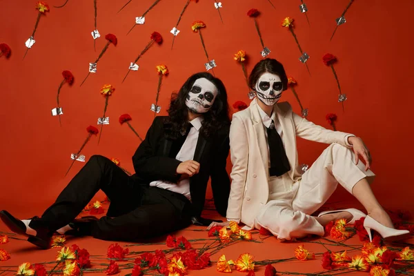 Пара в цукровому макіяжі черепа і елегантні костюми, що сидять на підлозі в червоній студії з квітами гвоздики — стокове фото