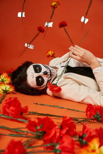 Жінка в макіяжі цукрового черепа і костюм лежить і дивиться на камеру біля гвоздик в червоній студії — стокове фото