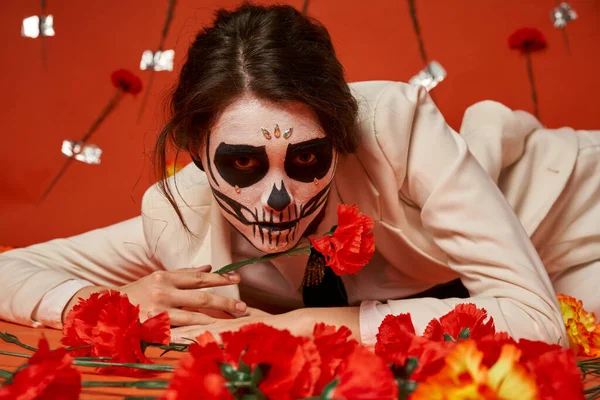 Elegante mujer en espeluznante catrina maquillaje acostado y mirando a la cámara cerca de las flores en el fondo rojo - foto de stock