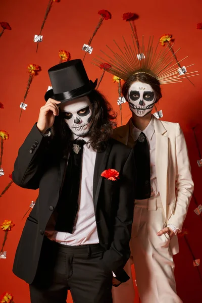 Elegantes Paar in Totenkopf-Make-up und festlichem Outfit auf rotem Hintergrund mit Blumen, Day of Dead Party — Stockfoto