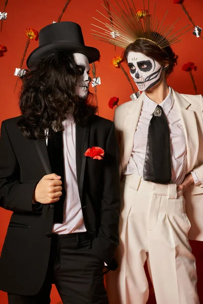 Dia de los muertos party, Paar in gruseliger Schminke schaut einander in rotem Studio mit Blumen an — Stockfoto
