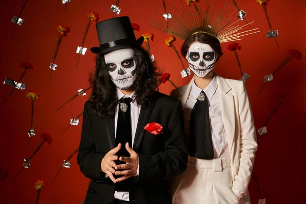 Día de los muertos fiesta, pareja en maquillaje de miedo mirando a la cámara en estudio rojo con claveles - foto de stock