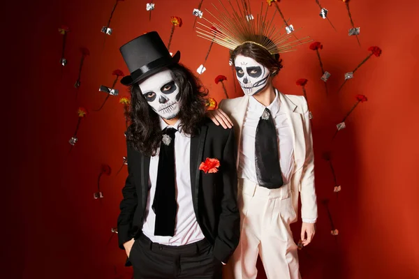 Elegante pareja en día de los muertos cráneo maquillaje mirando a cámara en rojo telón de fondo con flores - foto de stock