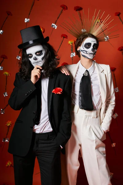 Couple dans le maquillage squelette effrayant et tenue festive sur fond rouge avec des fleurs, dia de los muertos — Photo de stock