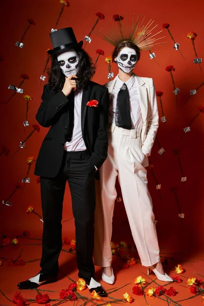 Piena lunghezza di festa elegante coppia in dia de los muertos trucco su sfondo rosso con decorazioni floreali — Foto stock